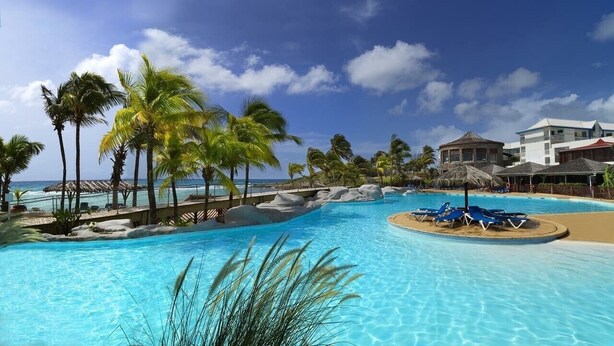 A large, clear swimming pool with palm trees and a beach view, surrounded by resort buildings. - Cozy single bedroom apartment with pool and private beach (Saint-François)