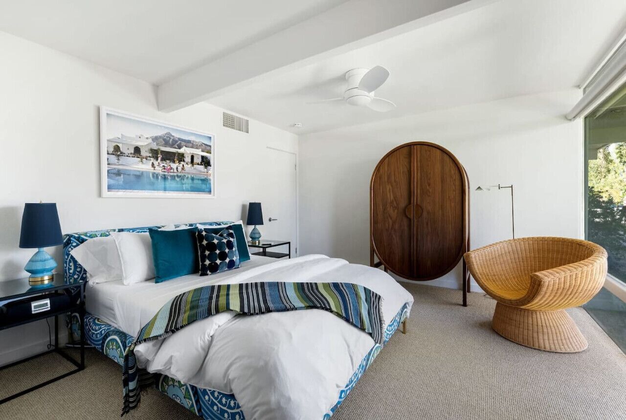 Une chambre à coucher avec un lit, une grande armoire en bois, une chaise en osier, un bureau avec une lampe et un tableau encadré au mur.