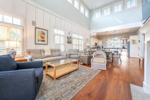 A spacious living area with a sofa, coffee table, and wicker chair, a kitchen in the background, and large windows.