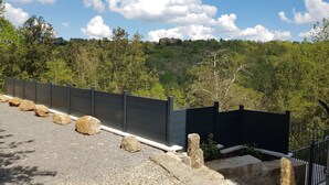 A stone wall with a metal fence, a gravel path, and a forested hillside in the background.