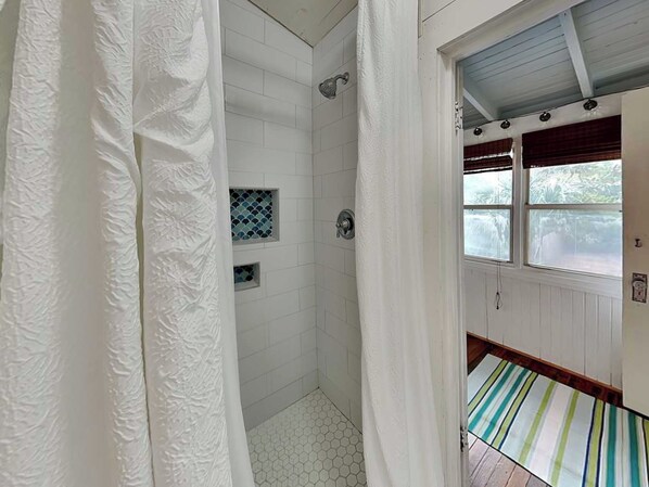 Une salle de bain avec des murs recouverts de carreaux blancs, une douche avec une porte en verre et une fenêtre avec des stores.