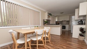 Une cuisine moderne avec des meubles blancs, une table à manger en bois et des chaises blanches, ainsi qu’une grande fenêtre avec des stores.