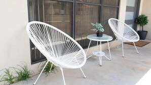 Deux chaises en osier blanc avec des pieds en métal, une petite table ronde et une plante en pot sur une terrasse en béton.