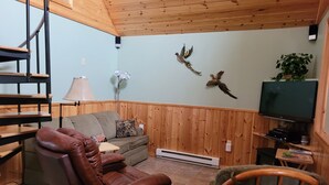 Un salón con un sillón de cuero marrón, un sofá gris, un mueble bajo de madera para televisor y un televisor montado en la pared.