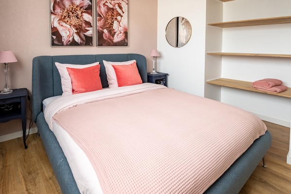 A neatly made bed with a pink blanket, a blue headboard, and two pillows. There are two bedside tables with lamps, and a wall-mounted shelf with a pink towel.