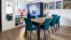 Un comedor con una mesa de madera y sillas color turquesa, una cocina al fondo y bloques de vidrio de colores en la pared.