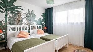 Una habitación con dos camas, un mural de plantas, una ventana con cortinas translúcidas y una alfombra colorida.