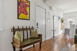 A wooden bench with patterned cushions, a white door, and a hallway leading to a kitchen area.