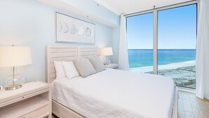 Ein Schlafzimmer mit einem großen Bett, zwei Nachttischen, einem Schreibtischchen, einem Wandbild und einem großen Fenster mit Blick auf den Strand.