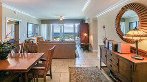 A spacious living area with a large sofa, dining table, and chairs. There is a view of the ocean through large windows.