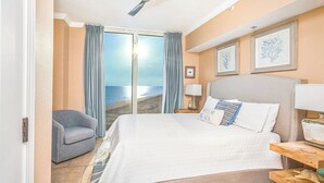 A hotel room with a bed, a chair, a ceiling fan, a nightstand, and a view of the ocean. - Sunrise views from your balcony! (Pensacola Beach)