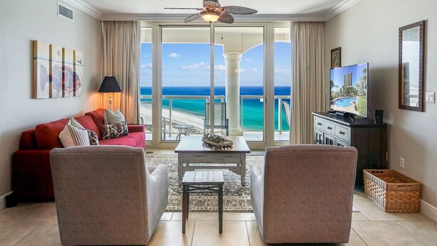 A living room with a red sofa, two armchairs, a coffee table, and a TV. The room has a large window with a view of the ocean.