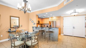 A dining area with a glass table and metal chairs, a kitchen with wooden cabinets, and a white door. - P3-2002 2B Gulf Front*Sunset Views! (Pensacola Beach)