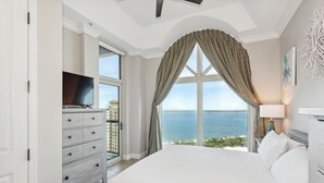 A bedroom with a large bed, a dresser, a TV, and a view of the sea. - Stunning Sound Views* Penthouse*P3-2106 (Pensacola Beach)