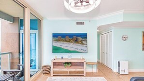 A living room with a large flat-screen TV, a wooden coffee table, a glass door leading to a balcony, and a ceiling fan. - Portofino Sky Home | Sound Views | Tower 4-0803 (Pensacola Beach)