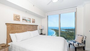 A bedroom with a large bed, a nightstand, a chair, and a view of the ocean. - P4-0902 2B Island View Oasis @ Portofino (Pensacola Beach)