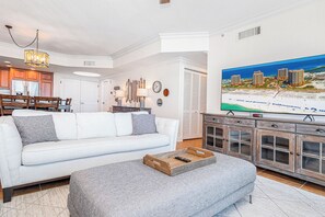 A living room with a white sofa, grey ottoman, wooden side table, and a flat-screen TV mounted on the wall.