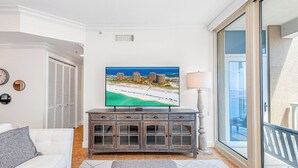 A living room with a television displaying a beach scene, a white sofa, a wooden cabinet, and a sliding glass door leading to a balcony. - P4-0902 2B Island View Oasis @ Portofino (Pensacola Beach)