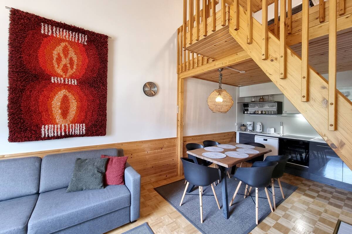 Una sala moderna con un sofá gris, un tapete rojo y naranja, una escalera de madera y una cocina con una mesa de comedor y sillas.