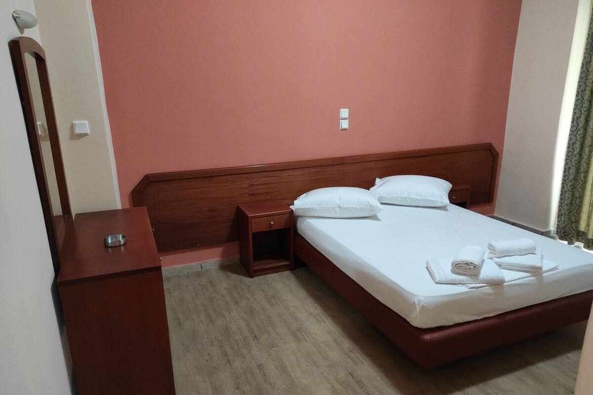 A neatly made bed with white linens and towels, a wooden nightstand with a drawer, a wooden headboard, and a mirror on the wall.