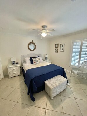A bedroom with a bed, nightstands, a ceiling fan, a window with blinds, and a wicker chair.