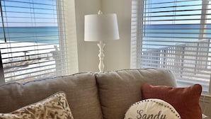 A living room with a beige sofa, patterned and solid-colored cushions, a white lamp, and a view of the beach through large windows. - "Sandy Toes" ON THE SAND! (Destin)