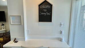A modern kitchen with a white island and two bar stools, a chalkboard sign above the island, and a window with blinds to the right. - "Sandy Toes" ON THE SAND! (Destin)