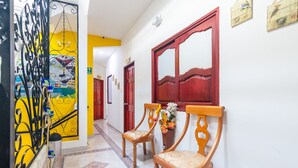 A hallway with two wooden chairs, a red door, and a yellow wall with a mural. - Hostal Kumbaya (Santa Marta)
