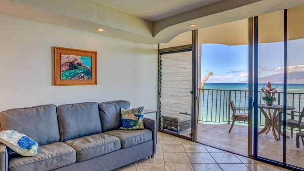 A balcony with a view of the ocean, a gray sofa with patterned cushions, and a framed picture on the wall.