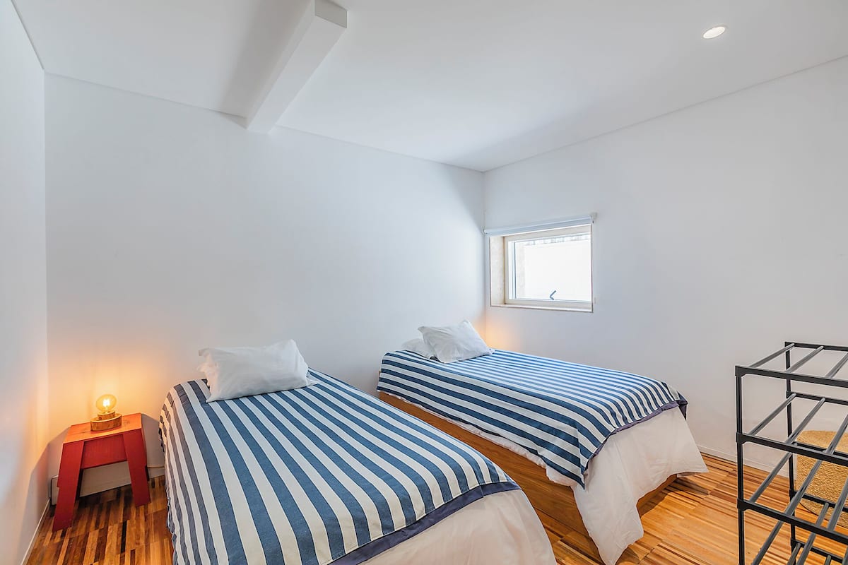 Une chambre avec deux lits, une petite fenêtre, une table de chevet et un étagère en métal.