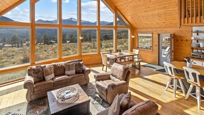 Amplia habitación con techo alto y ventanas grandes que ofrecen vistas a la montaña. Cuenta con un rincón de estar acogedor con sofá y sillones, además de una mesa de comedor con sillas.