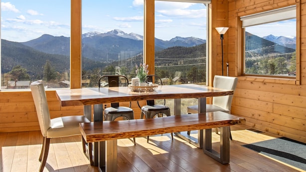 Interior de madera con una mesa de comedor, sillas y un banco. Un ventanal ofrece vistas a montañas y un valle.
