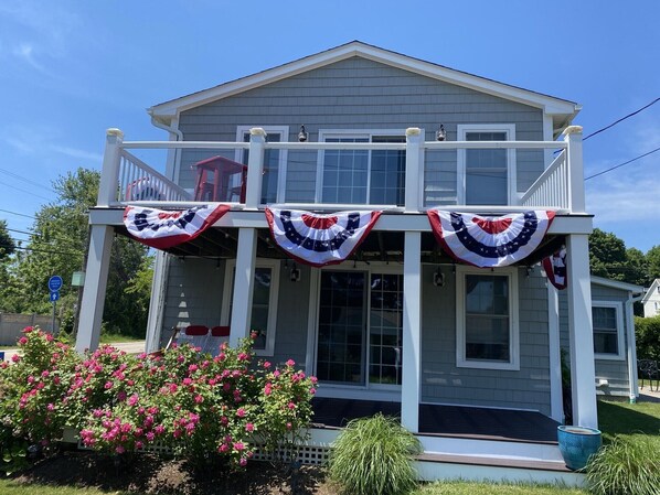 A two-story house with a balcony, a porch with American flags, and a garden with pink flowers.