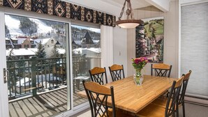 Un coin repas avec une table et des chaises en bois, un vase rempli de fleurs et, à travers une porte vitrée, une vue sur un village enneigé.