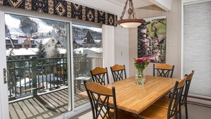 Un coin repas avec une table et des chaises en bois, un vase rempli de fleurs et, à travers une porte vitrée, une vue sur un village enneigé.