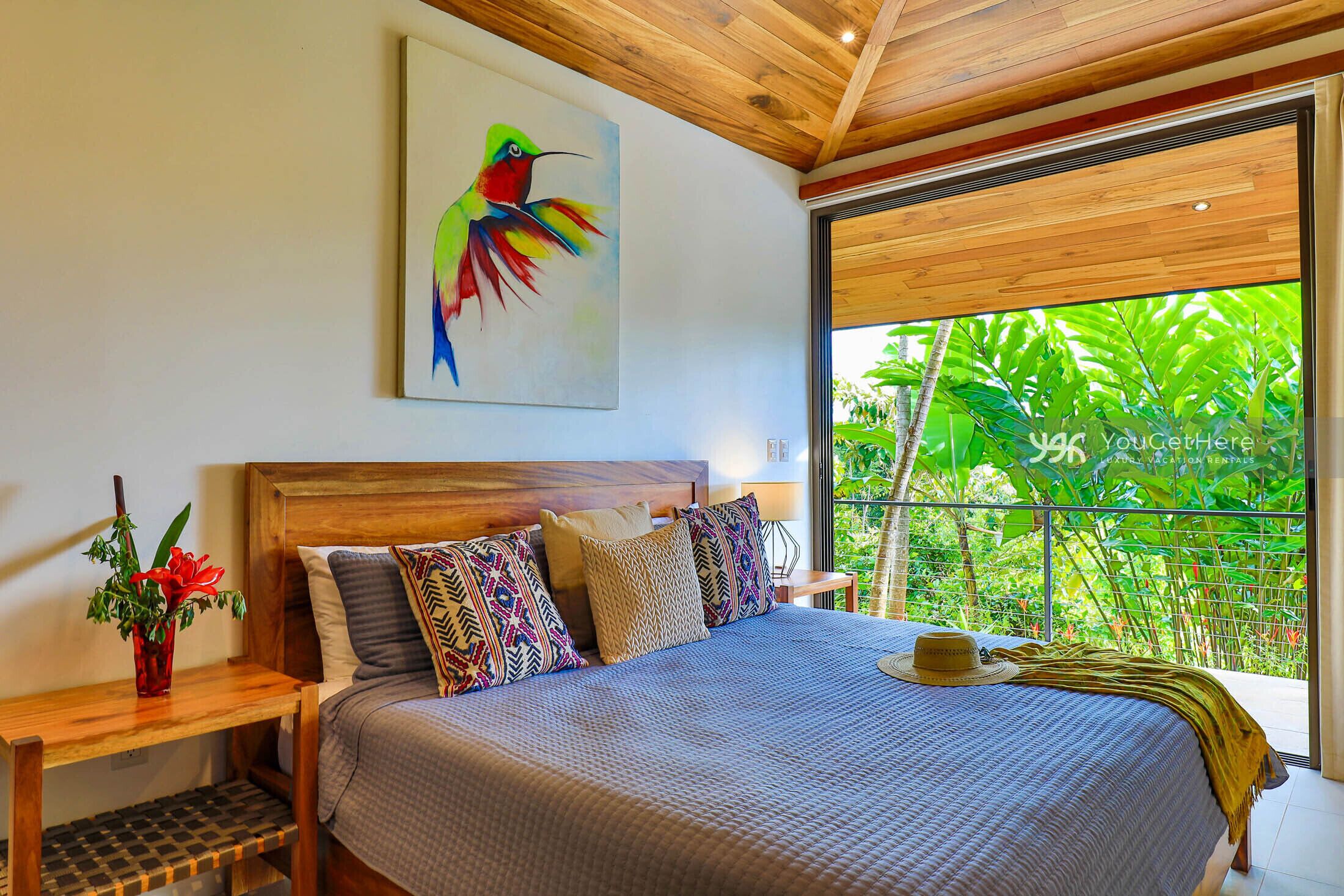 Un dormitorio con una cama grande, cabecera de madera, un cuadro de un pájaro, una mesita de noche con un jarrón de flores y vistas a la vegetación a través de una ventana.
