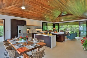 A modern kitchen with a central island, dining area with wicker chairs, and large windows offering a view of greenery.