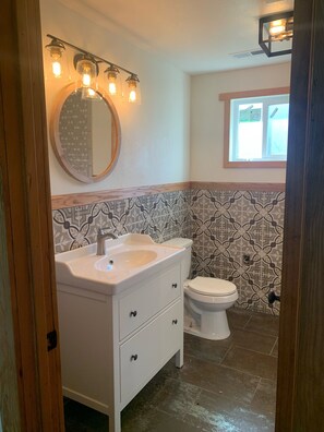 Une salle de bain avec un meuble-lavabo blanc, un miroir rond et des toilettes. Des luminaires décoratifs sont placés au-dessus du miroir et une fenêtre se trouve au-dessus des toilettes.