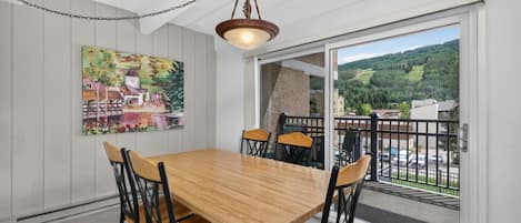 Un comedor con una mesa y sillas de madera, una lámpara colgante, un cuadro en la pared y vistas a un paisaje verde a través de una puerta de vidrio.