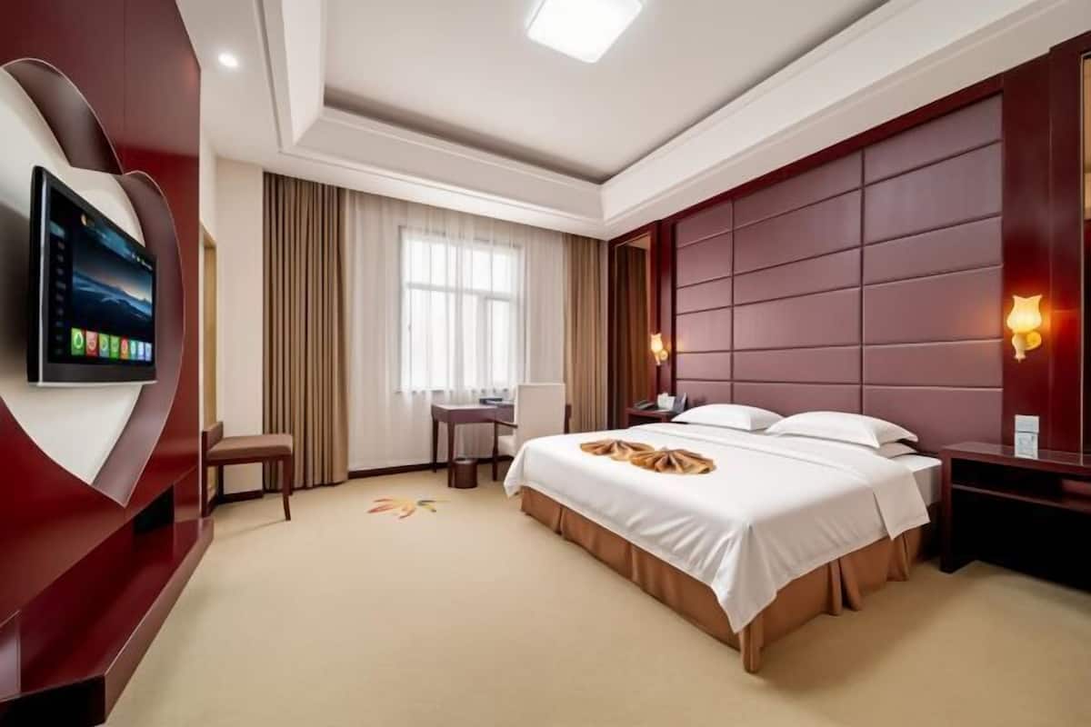 Une chambre d’hôtel avec un grand lit, un bureau, une chaise, un téléviseur fixé sur un mur incurvé et une fenêtre avec des rideaux.
