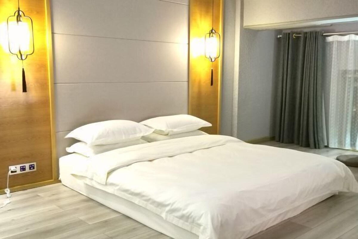 A hotel room with a bed, two wall-mounted light fixtures, a window with curtains, and a power outlet on the wall.