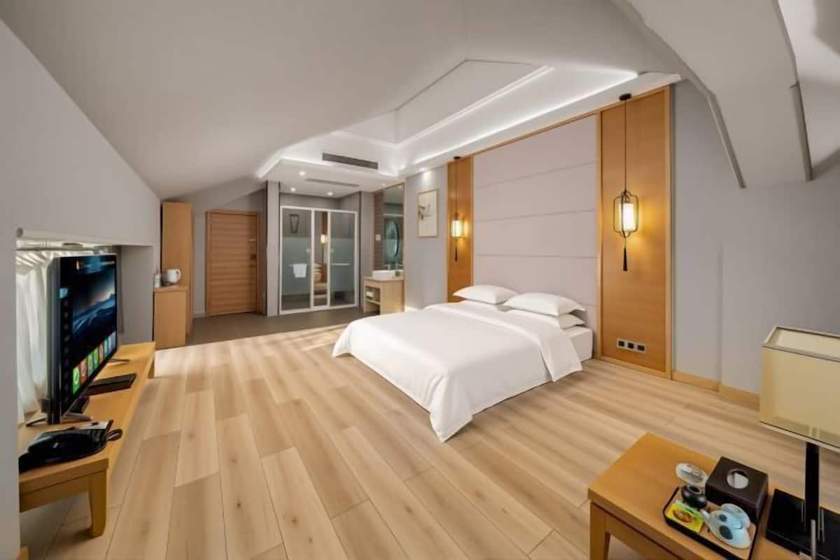 Une chambre d’hôtel moderne avec un plancher en bois, un lit, une télévision, un bureau et une salle de bain visible à travers une porte en verre.