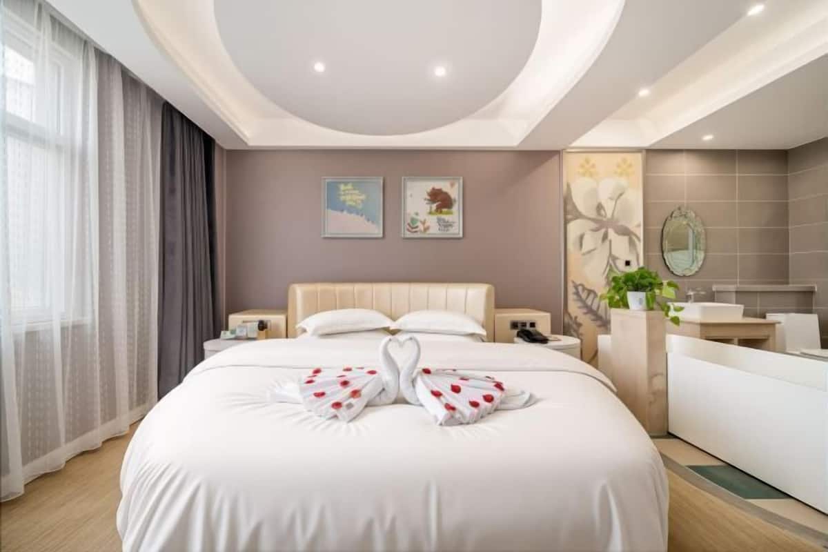 Une chambre d’hôtel avec un grand lit, une table de chevet, une commode, une salle de bain avec un lavabo et un miroir, et une fenêtre avec des rideaux.