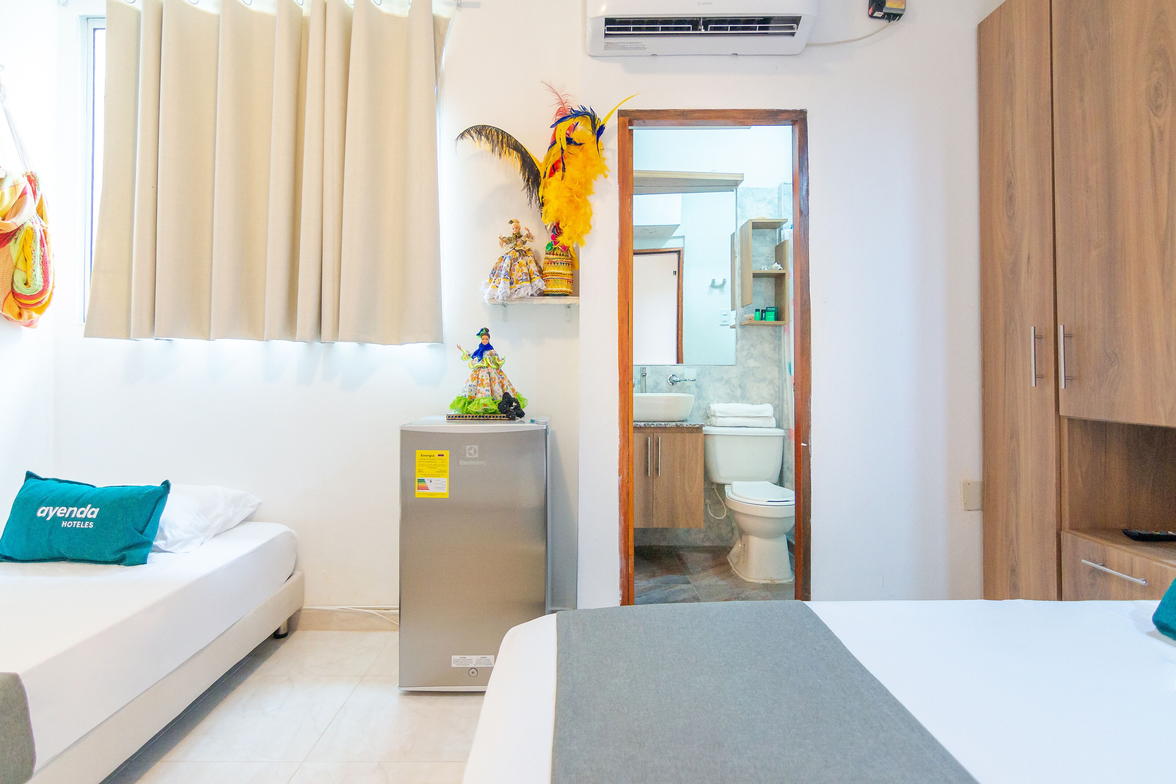A hotel room with a bed, a small refrigerator, a wooden wardrobe, and a bathroom visible through an open door.