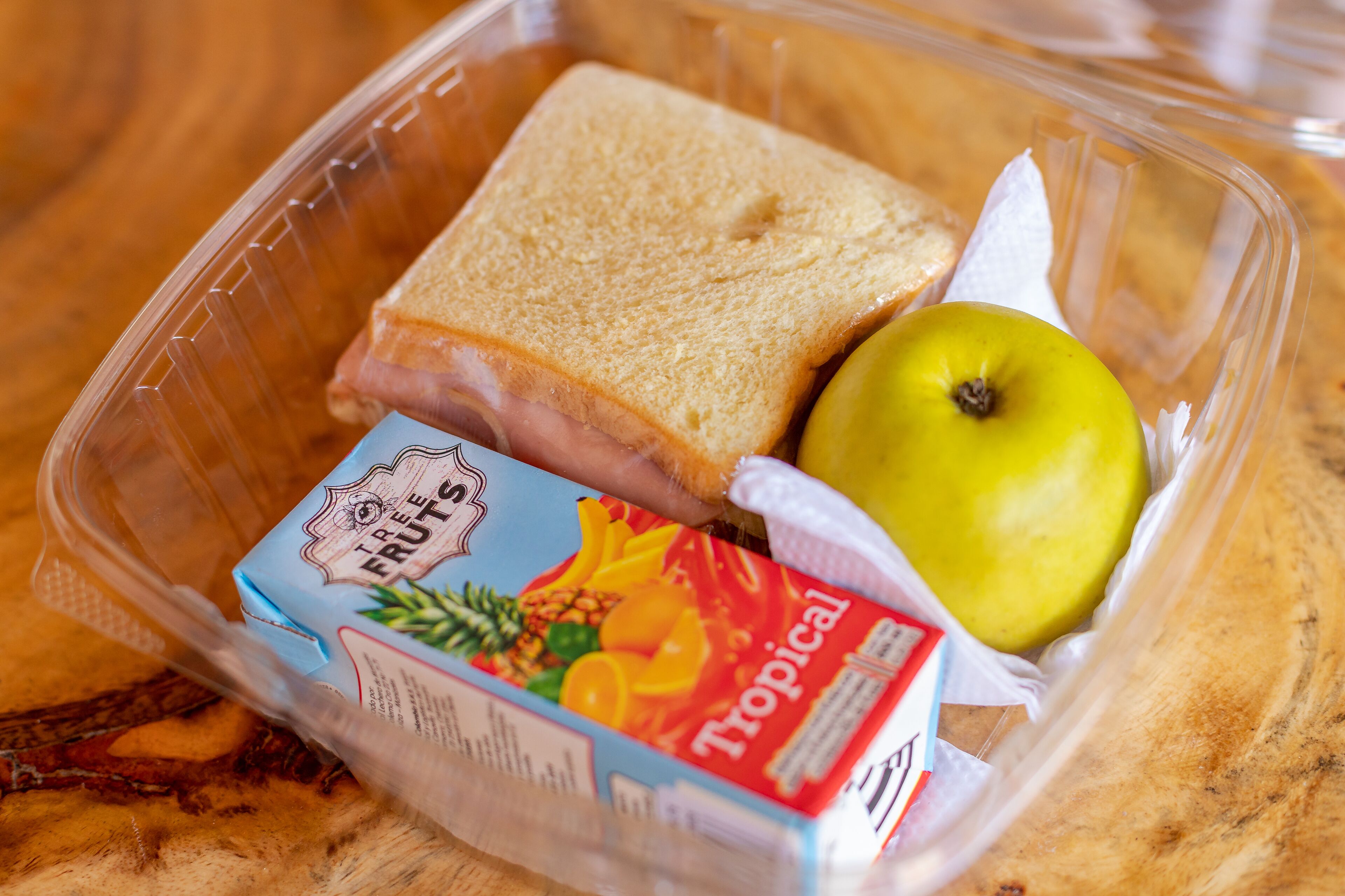 A clear plastic container with a slice of white bread, a green apple, and a carton of tropical fruit juice.