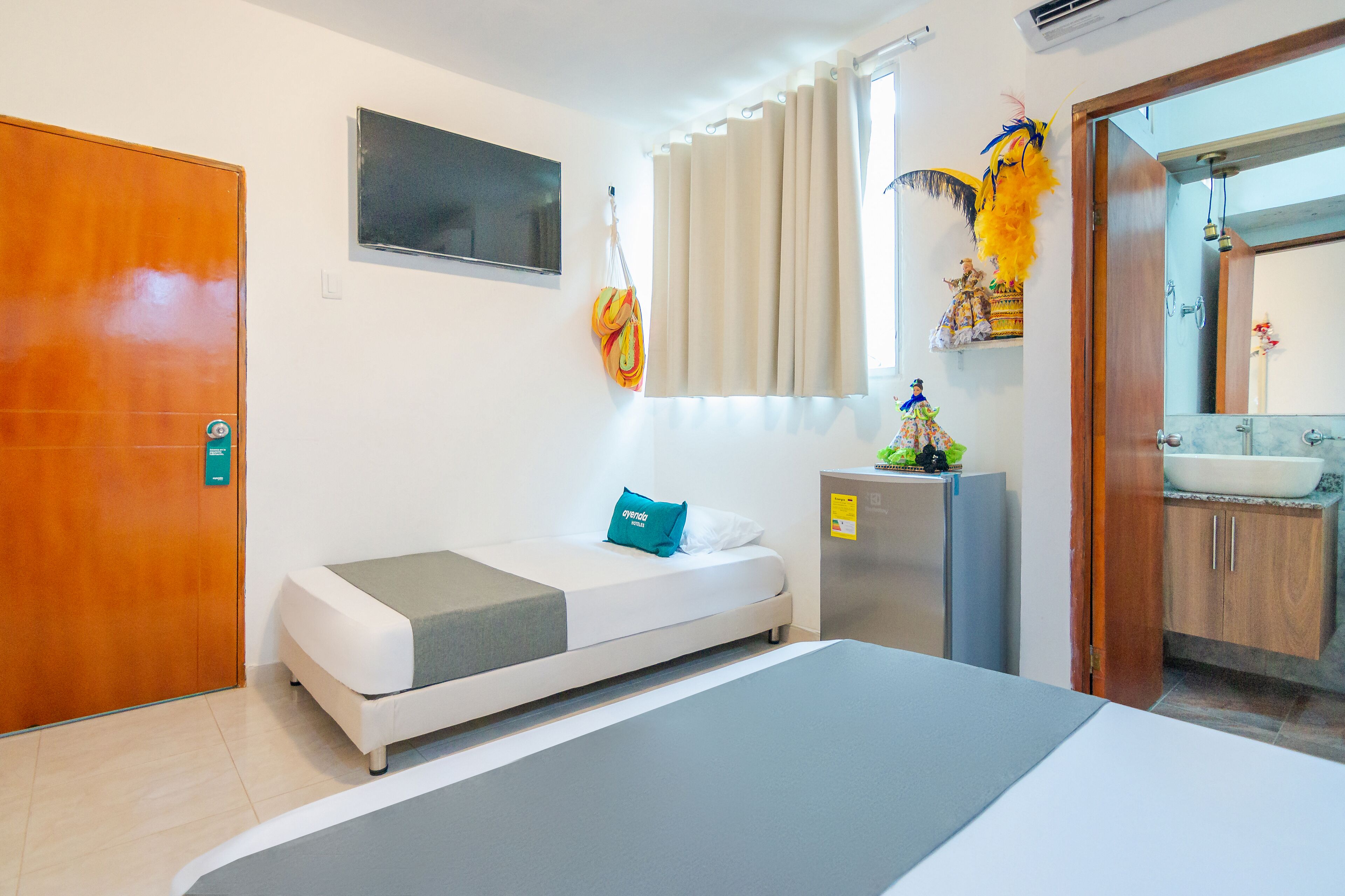 Quarto de hotel com uma cama, uma escrivaninha, um espelho, uma pia e uma porta.