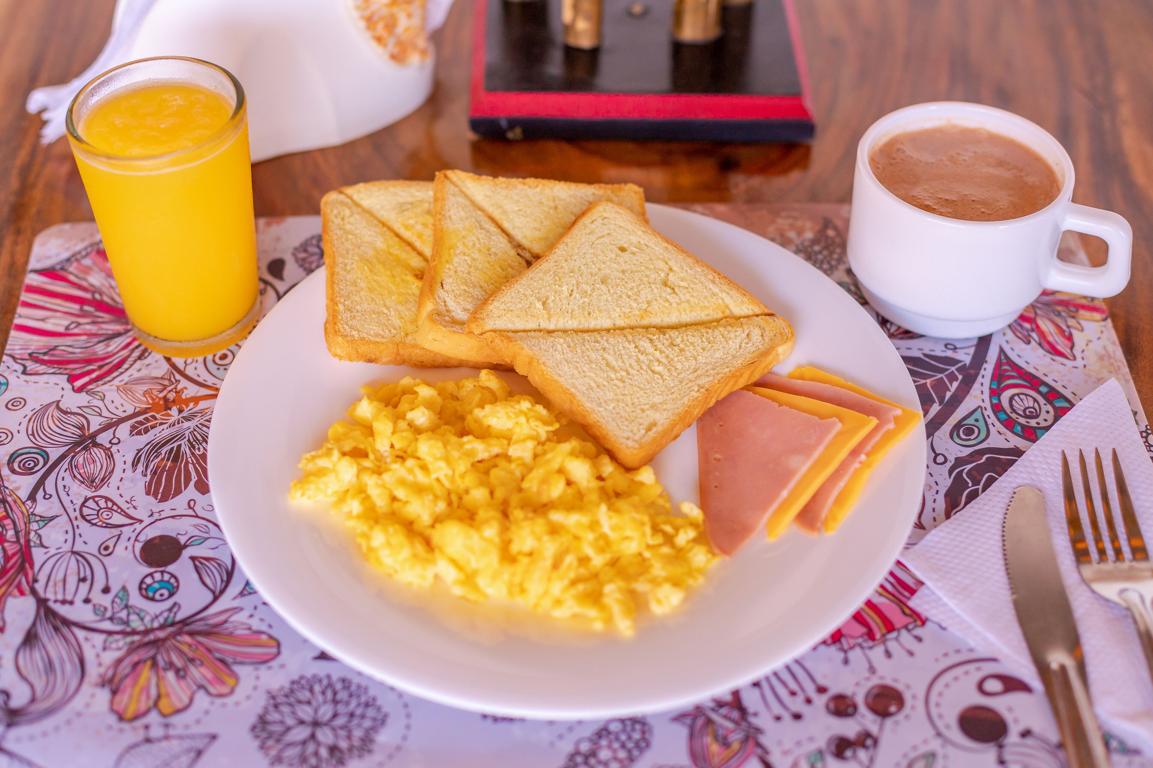Un plato de desayuno con huevos revueltos, jamón y tostadas, un vaso de jugo de naranja y una taza de café sobre un mantel estampado.