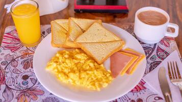 Un plato de desayuno con huevos revueltos, jamón y tostadas, un vaso de jugo de naranja y una taza de café sobre un mantel estampado.