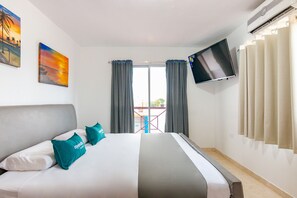 Ett hotellrum med en säng, två tavlor, en tv och en balkong med gardiner.