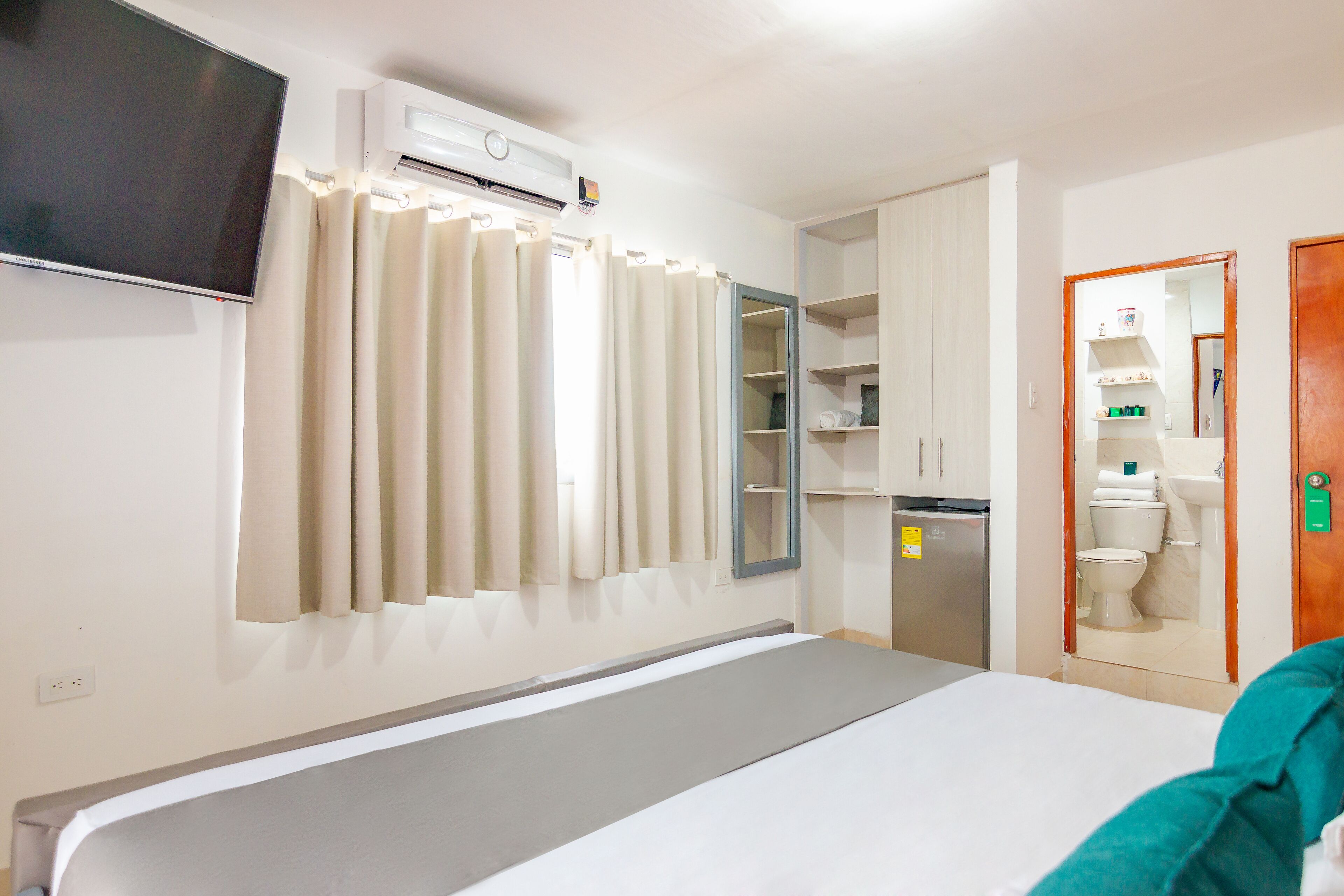 Une chambre d’hôtel comprenant un lit, un téléviseur fixé au mur, une armoire, un miroir et une salle de bain avec des toilettes et un lavabo.